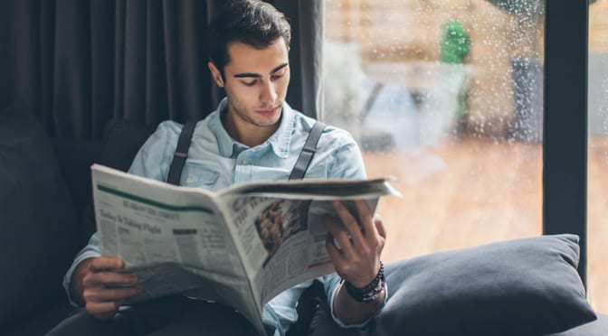 man sitting down on a couch reading a newspaper